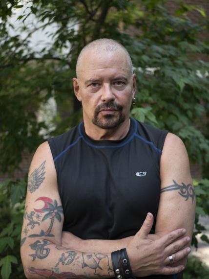 Sky Gilbert standing in front of trees, his arms folded, showing tattoos. 